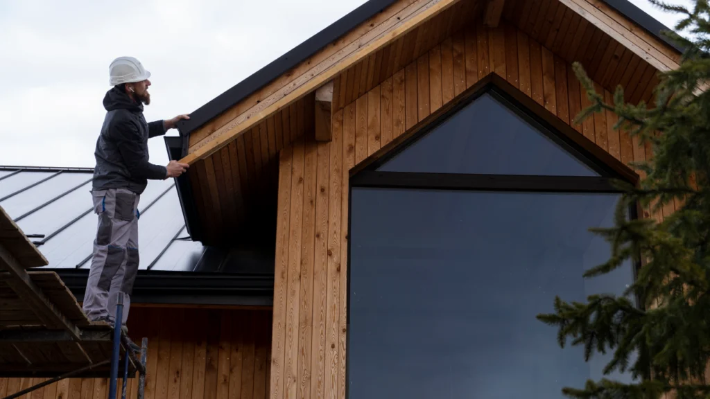 Professional gutter cleaning worker on ladder removing debris from gutters on modern wood-sided house with metal roofing.RetryClaude can make mistakes. Please double-check responses.