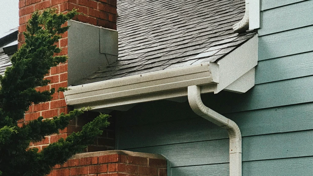 House exterior showing white gutters and downspout on blue-green siding with brick chimney and asphalt shingle roof.